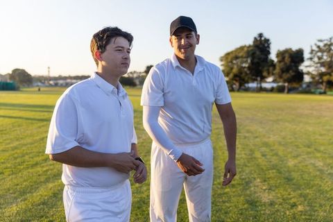 Two teammates in white cricket uniforms standing on sunlit outfield during training