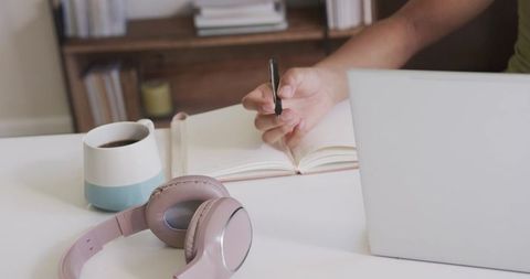 Woman writing in notebook at minimal home desk with laptop, coffee mug and headphones