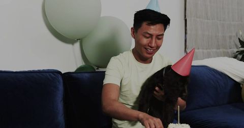 Smiling man celebrating birthday with dog on couch wearing party hats and cutting cake