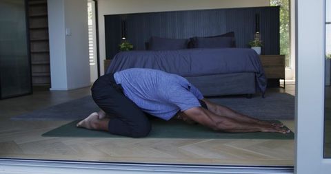 Man practicing child's pose on home yoga mat in modern minimalist bedroom for wellness