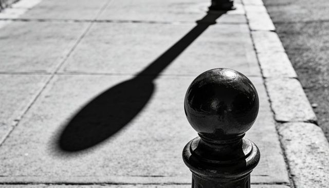 Black bollard casting long lamppost shadow on urban sidewalk high-contrast monochrome