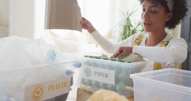 Woman organizing recyclable materials into separate bins at home. She is putting plastic and paper waste into clearly labeled, transparent containers. This photo can be used to promote eco-friendly practices, waste management methods, and environmental awareness.