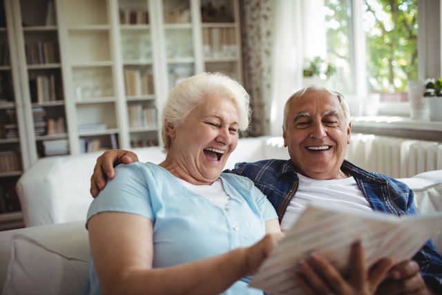 Senior couple sitting on a couch in a cozy living room, looking at a photo album and smiling. This image can be used for themes related to family, memories, retirement, and elderly lifestyle. Ideal for advertisements, articles, and blogs focusing on senior living, family bonding, and nostalgic moments.