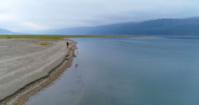 Person walking alone by the serene lakeshore, creating a peaceful and tranquil scene. Ideal for use in themes of solitude, relaxation, nature appreciation, and outdoor activities. Suitable for blog posts, travel websites, and stress-relief visualizations.