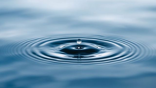 Balancing crystal water droplet forming concentric ripples on calm blue surface