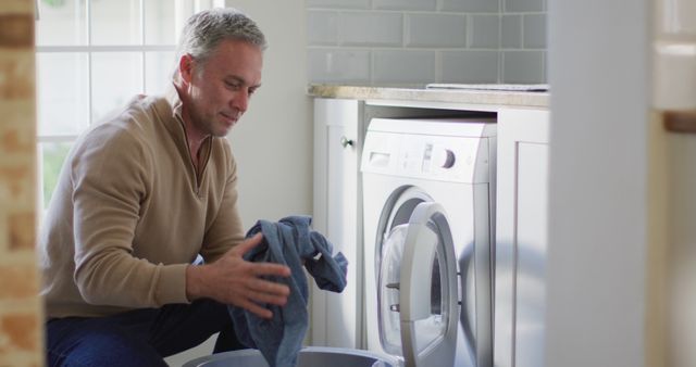 Mature man doing laundry at home in a modern washroom. Ideal for use in content related to daily chores, household responsibilities, home appliances, and domestic lifestyle. Can be used in ads for laundry products, washing machines, or home cleaning services.