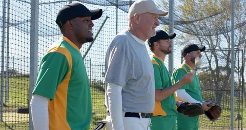 Multiracial adult baseball teammates standing by backstop watching game and talking