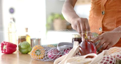 Person placing fresh produce into reusable bag in modern kitchen