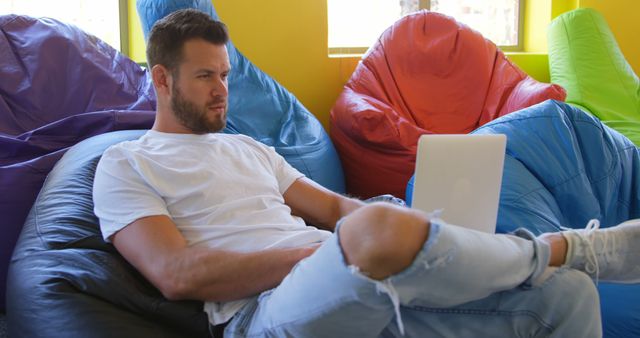Young man sitting on black bean bag chair, working on laptop in a modern, colorful workspace with bright yellow, red, green, and blue bean bags around. Ideal for illustrating remote work, tech industry job, modern office environment, and casual professional settings.