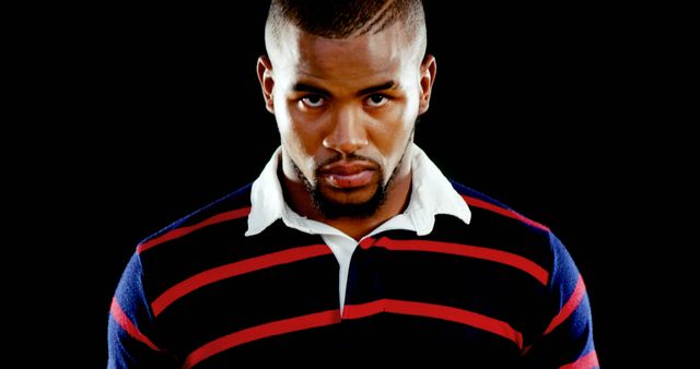 Young African American man wearing striped sweater, gazing directly at camera with confident expression. Black background focuses attention on subject. Ideal for topics on self-confidence, youth culture, fashion apparel, and personal development.