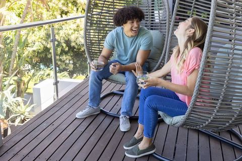 Sunlit balcony conversation: young couple laughing in woven egg chairs with drinks