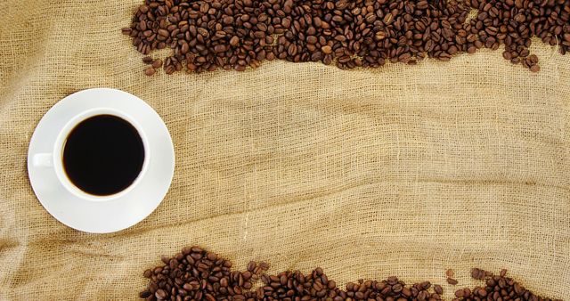 A cup of black coffee sits on a burlap surface surrounded by scattered coffee beans, with copy space. Ideal for themes related to morning routines, coffee culture, or the beverage industry.