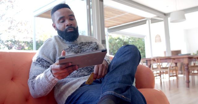 Man sitting on a couch using a tablet, likely engaged in online shopping. He is wearing a sweater and jeans, situated in a bright and modern living room. Perfect for illustrating online retail, digital commerce, or modern lifestyle themes.