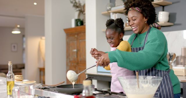 Mother and daughter smiling while making pancakes in modern kitchen. Shows family bonding, joy, and teaching moments. Ideal for promoting family-oriented products, cooking tutorials, or parenting blogs.
