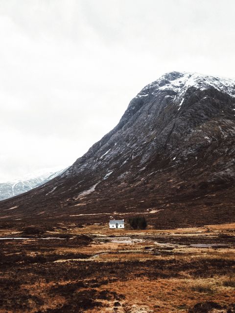 An isolated white cottage stands out against the rugged terrain of the Scottish Highlands with a majestic mountain backdrop. The surrounding landscape is vast and wild, providing a sense of tranquility and solitude. Ideal for travel blogs, adventure tourism advertisements, or serene landscape artwork.