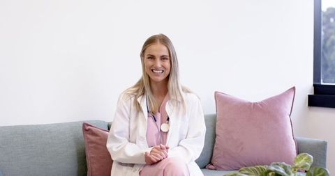 Female Clinician Sitting on Sofa Wearing Pink Scrubs and White Lab Coat Smiling Calmly