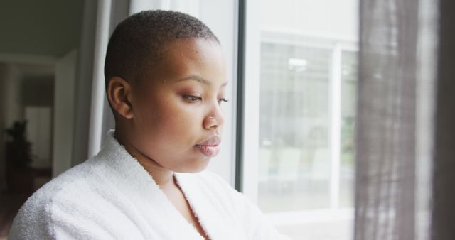 Thoughtful african american plus size woman wearing robe, looking through window. domestic lifestyle, enjoying leisure time at home.