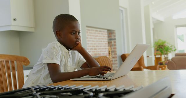 Young boy sitting at kitchen counter using laptop for online education or e-learning. Ideal for use in articles and advertisements focused on online schooling, children's use of technology, and distance learning. Great for featuring the home learning environment and modern education tools.