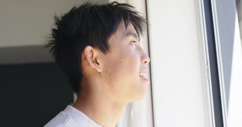 Smiling asian man gazing out bright window at home, sunlit profile portrait closeup