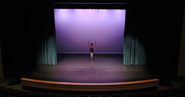 A young Caucasian girl is standing on stage with her arms raised in a theater, with copy space. Her pose suggests she may be a dancer or performer practicing or taking a bow after a performance.