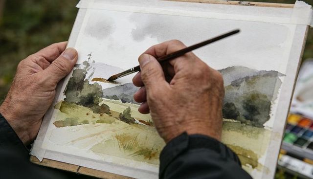 Painting watercolor landscape plein air: artist brushing field, hills, trees on paper