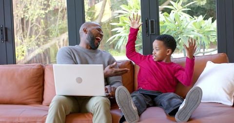 Laughing African American father and son bonding on leather couch with laptop and garden