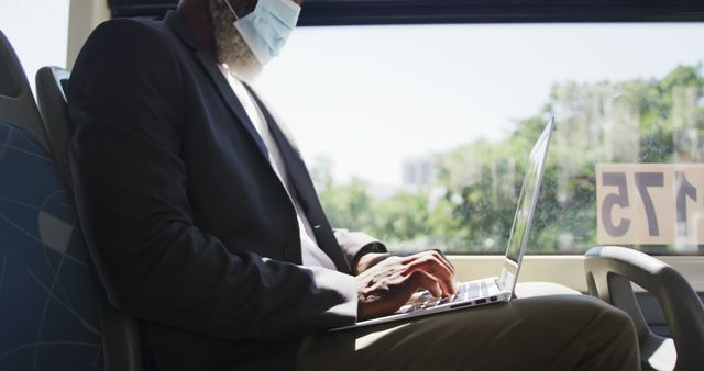 Business professional wearing face mask traveling on public bus, working on laptop with focused expression. Ideal for use in reports or articles about remote work, commuting, safety measures in public transportation, and modern ways to balance work and travel.