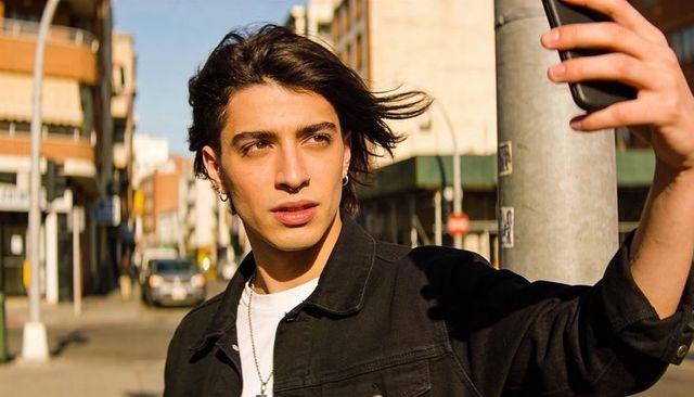 Young man taking selfie on sunlit urban corner, windblown hair and silver jewelry