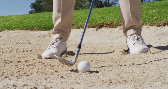 This is a close-up view of a golfer's feet and club preparing to hit a ball from a sand trap. Ideal for use in articles or advertisements related to golf techniques, golfing equipment, sports events, and golf courses.
