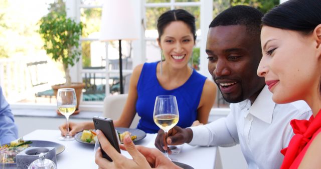 Group of friends enjoying lunch at an outdoor restaurant, looking at a mobile phone while mingling and drinking wine. Perfect for illustrating social gatherings, modern lifestyle, outdoor dining, and technology usage in social settings.