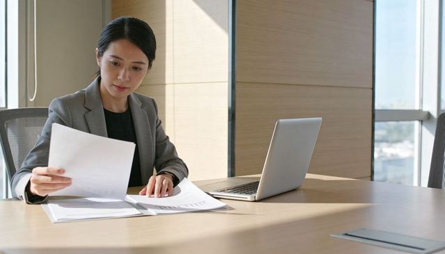 Asian businesswoman reviewing documents in bright modern conference room with laptop