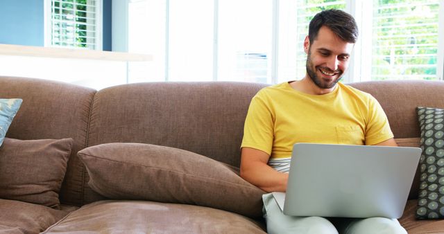 Young man in yellow shirt sitting on a brown couch, happily using a laptop. Light-flooded living room with modern decor. Ideal for illustrating remote work environments, home offices, or casual technology usage from home.