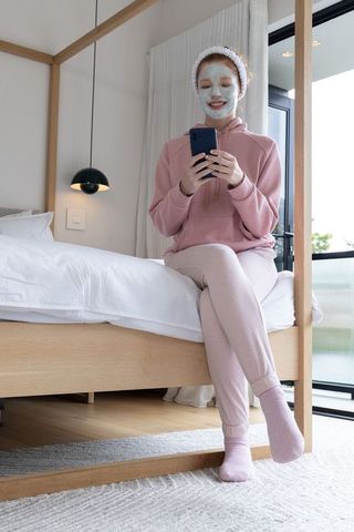 Young woman relaxing on bed wearing face mask and pink loungewear holding smartphone
