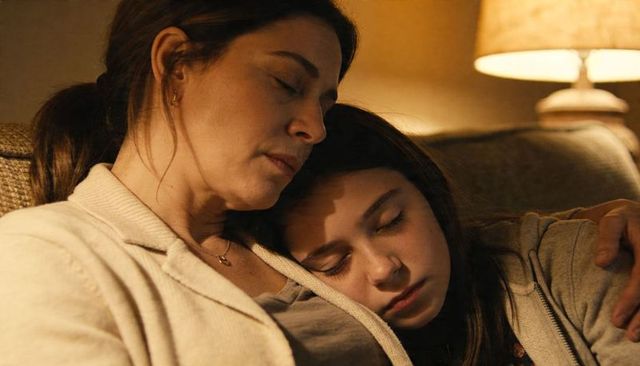 Mother cradling teenage daughter sleeping on sofa under warm lamp glow, cozy family bond