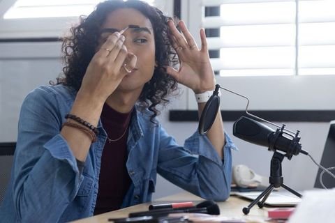 Non-binary beauty creator applying eyebrow makeup during live stream with microphone