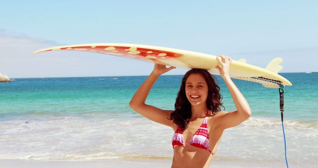 Showing a woman smiling while holding a surfboard above her head on a sunny beach, this image captures the essence of summer fun and outdoor activities. Ideal for promoting beach vacations, water sports, travel destinations, or lifestyle brands. Can be used to depict concepts like freedom, happiness, and enjoyment.