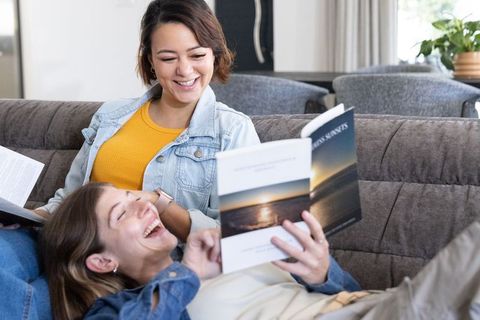 Relaxing couple laughing and reading while reclining on gray sofa in cozy sunlit living room
