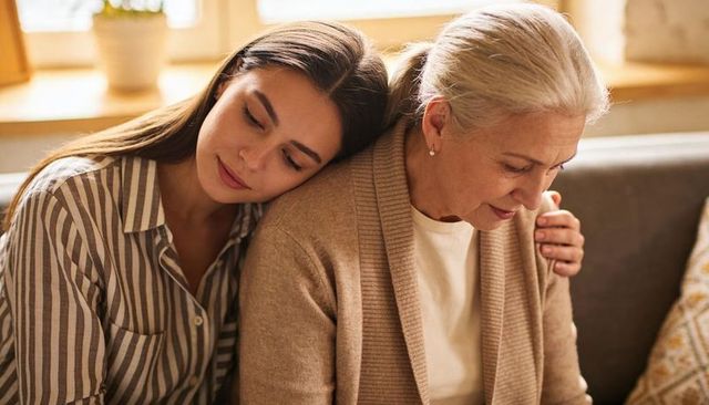 Adult daughter leaning on elder mother sharing warm hug on sunlit cozy couch