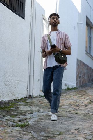 Young Asian man walking cobblestone alley listening to music and using smartphone