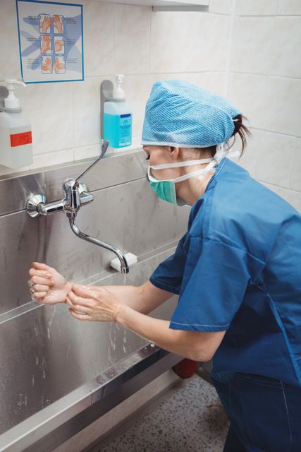 Female surgeon in blue scrubs washing hands at a hospital sink. She is wearing a surgical cap and mask, following hygiene protocols. Useful for topics related to healthcare, hygiene, medical procedures, and infection control.