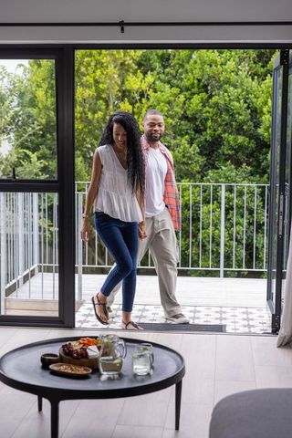 Diverse couple entering modern living room through sliding doors holding hands on balcony