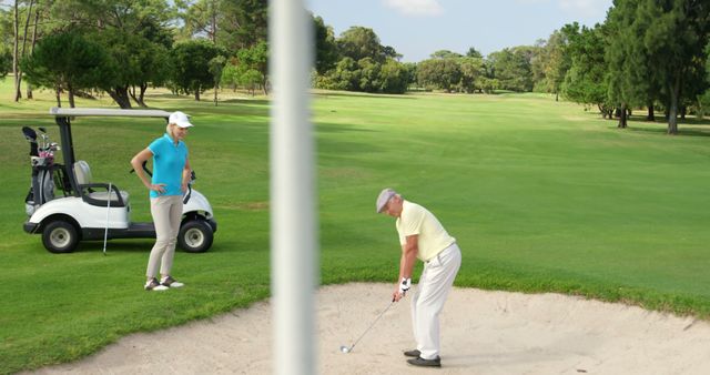 Elderly man playing golf, focused on hitting ball from sand bunker while female golfer watches. Ideal for advertisements related to golfing, active seniors, sports, or retirement activities. Perfect for magazines, websites, and promotions focusing on sportsmanship, teamwork, and outdoor activities.