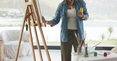 Creative woman painting on canvas at wooden easel in home studio with waterfront view