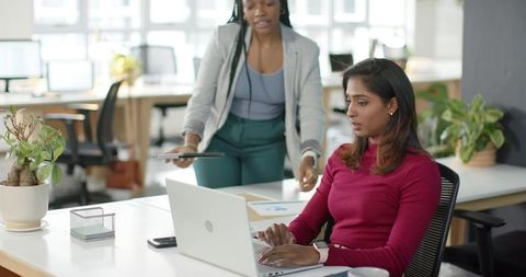 Multicultural female colleagues collaborating at open-plan office using laptop and tablet