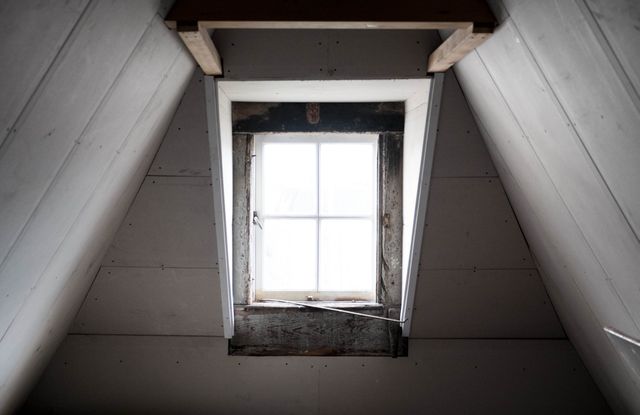 Sunlight is shining through a lone window in an empty attic space. The rustic design with wooden beams and aged wood creates a vintage atmosphere, perfect for concepts related to isolation, quiet spaces, historical settings, architecture, and minimalism. This can be used in articles, blogs, and advertisements focusing on home improvement, interior design, and serene environments.