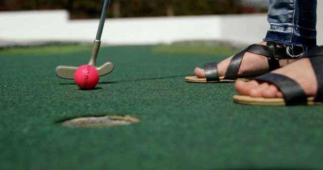 An individual is playing mini-golf on a green course with focus on feet in open toe sandals and a putter. The scene captures the moment just before hitting a pink mini-golf ball towards the hole. Ideal for depicting leisure activities, casual sports events, lifestyle concepts, or promotional content for resorts and golf courses.