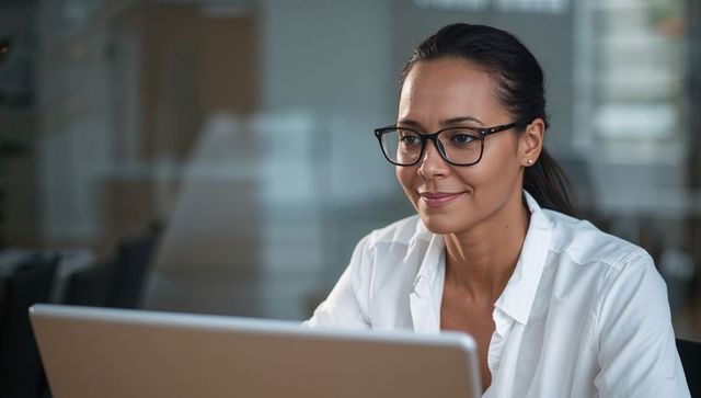 Focused professional woman in glasses working on laptop in office with glass reflections