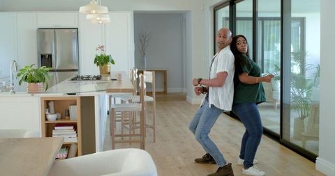 Diverse couple dancing back-to-back in sunlit modern kitchen while holding smartphones