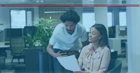 Reviewing document together in open-plan office, woman holding paper, man pointing tablet