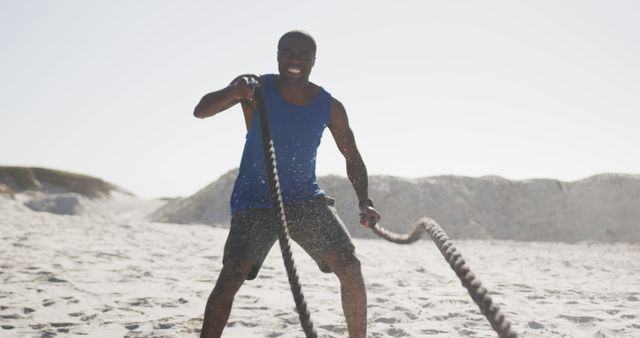 Smiling african american man exercising with battling ropes outdoors on beach. fitness, healthy and active lifestyle concept.